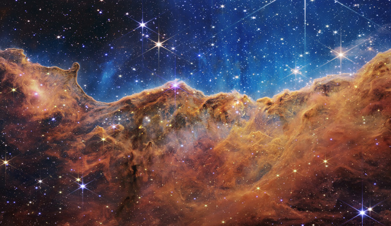 An undulating, translucent star-forming region in the Carina Nebula is shown in this Webb image, hued in ambers and blues; foreground stars with diffraction spikes can be seen, as can a speckling of background points of light through the cloudy nebula. Credit: NASA's Goddard Space Flight Center